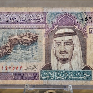 May include: A five Saudi Riyal banknote. The bill features an image of a man in traditional headdress, a seascape with boats, and Arabic text. The bill is primarily purple, blue, and green.