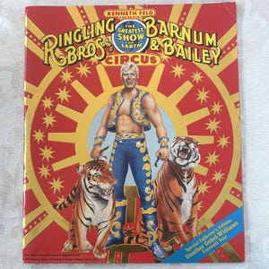 May include: A souvenir program cover for Ringling Bros. and Barnum & Bailey Circus. The cover features a muscular man in a blue and gold outfit with two tigers. The background is red with gold accents and the text "The Greatest Show on Earth".