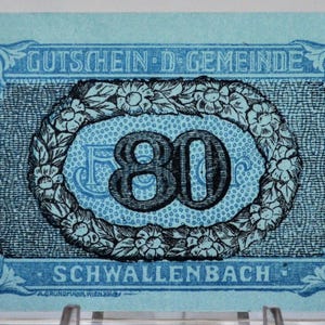 May include: A vintage blue and white document from 1920, featuring the text "GUTSCHEIN-D-GEMEINDE" and "SCHWALLENBACH". The central design includes the number "80" within a floral wreath. The corners have decorative scrollwork.