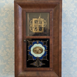 May include: A rectangular wall clock with a dark brown wooden frame. The upper section features a visible brass gear mechanism with gold hands. The lower section has a black glass panel with blue and gold accents surrounding a central oval illustration.