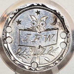 May include: A silver circular emblem featuring an owl, stars, and a crescent moon. The emblem has the initials "JTW" on an open book. The outer edge has a decorative border with small holes.