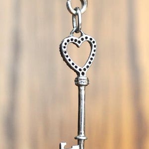 May include: A silver heart-shaped key pendant with a decorative design. The key has a small loop at the top for attaching to a chain or bracelet.