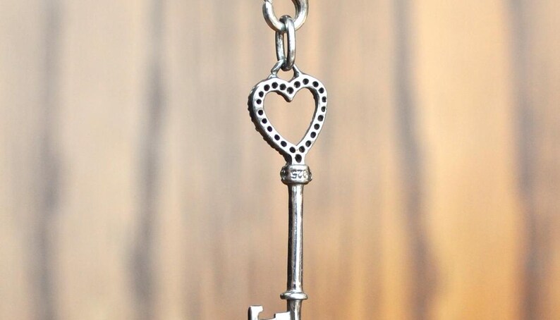 May include: A silver heart-shaped key pendant with a decorative design. The key has a small loop at the top for attaching to a chain or bracelet.