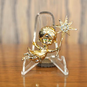 May include: A gold-toned brooch featuring a fairy with green eyes holding a star-shaped wand with clear rhinestones. The fairy is wearing a flowing dress and has a whimsical expression.
