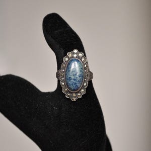 May include: An oval-shaped silver ring with a large blue stone and a halo of smaller, clear stones. The ring is displayed on a black velvet hand, showcasing its intricate design and vintage style.