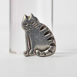 May include: A silver-toned metal pin shaped like a sitting cat with engraved facial features and striped markings along its back and tail. The cat is depicted in a seated position with its tail curled around its side, featuring a smooth metallic finish.