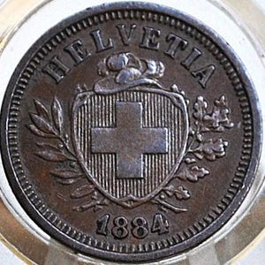May include: A close-up of a bronze coin with the word "HELVETIA" arched across the top. The coin features a shield with a cross and the year "1884" at the bottom. The coin is encased in a clear, circular holder.