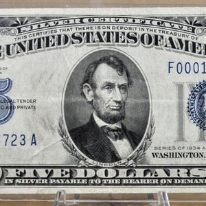 May include: A vintage 1934 United States five-dollar silver certificate. The bill features a portrait of Abraham Lincoln and the text "FIVE DOLLARS IN SILVER PAYABLE TO THE BEARER ON DEMAND". The bill is encased in a clear protective sleeve.