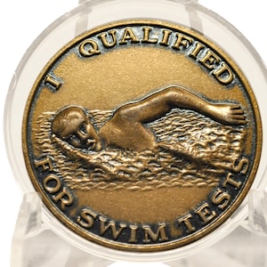 May include: A circular bronze-toned medal featuring a raised relief of a person swimming in water. The text "I QUALIFIED" is arched across the top, and "FOR SWIM TESTS" is arched across the bottom in raised, dark-colored capital letters.