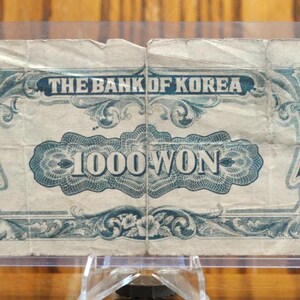 May include: A vintage 1000 Won banknote from The Bank of Korea. The note features intricate blue and white floral designs and the denomination is printed in bold blue lettering.
