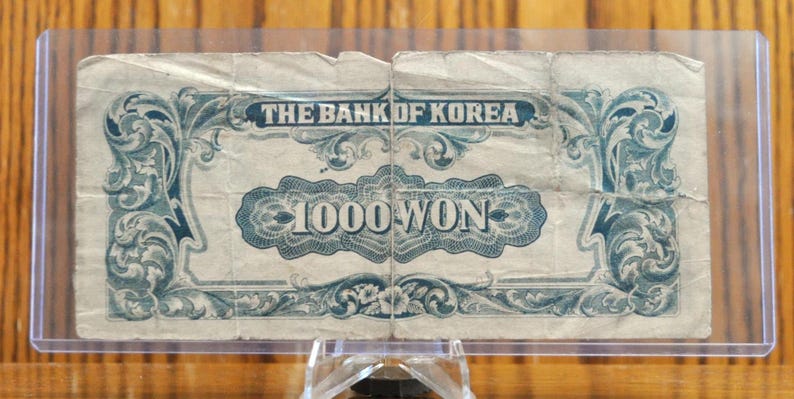 May include: A vintage 1000 Won banknote from The Bank of Korea. The note features intricate blue and white floral designs and the denomination is printed in bold blue lettering.