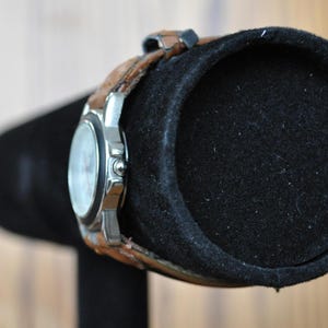 May include: A silver wristwatch with a white face and brown leather band is displayed on a black watch stand.