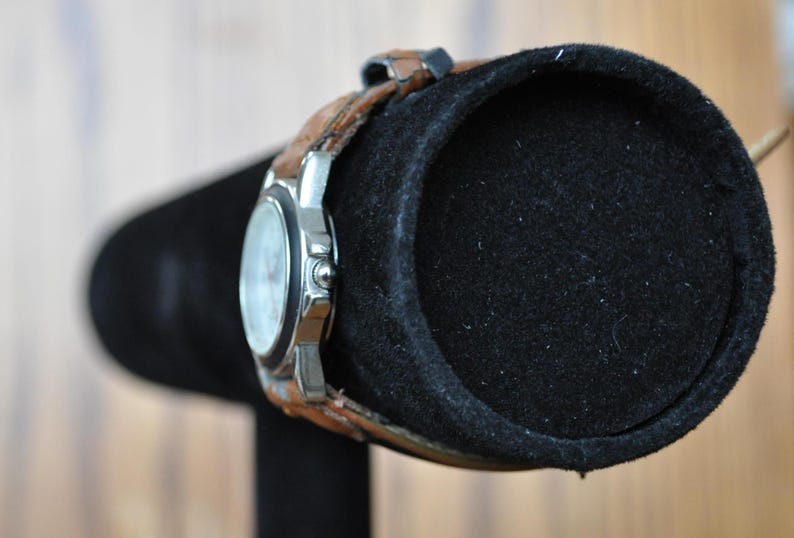 May include: A silver wristwatch with a white face and brown leather band is displayed on a black watch stand.