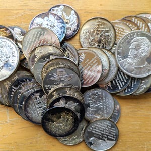 Puede incluir: Una colección de monedas de plata con varios diseños e inscripciones. Algunas monedas presentan figuras históricas, mientras que otras muestran imágenes y textos religiosos. Las monedas están dispuestas sobre una superficie de madera.