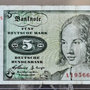 May include: A vintage five Deutsche Mark banknote. The bill features a portrait of a woman and the text "Banknote Fünf Deutsche Mark". The serial number "A 1956676 S" is printed in red. The bill is approximately 14 cm long.