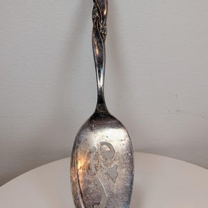 Puede incluir: Cuchara de servir plateada con un mango decorativo y un diseño floral. El mango tiene un diseño retorcido con detalles florales. El cuenco de la cuchara presenta un patrón floral recortado. Un utensilio de cocina vintage.