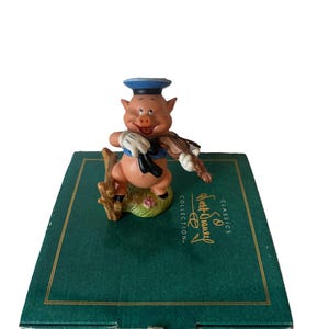 Könnte beinhalten: Figur eines lächelnden Schweins mit Matrosenmütze und Handschuhen, das Geige spielt. Das Schwein steht auf einem Grasbüschel mit Blumen. Die Figur steht auf einer grünen Schachtel mit dem Text "Walt Disney Classics Collection."
