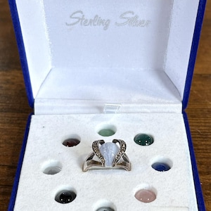 May include: A sterling silver ring with a light blue teardrop stone and small accent stones, presented in a blue velvet box with seven additional loose cabochon gemstones in various colors and a label indicating Size 7.