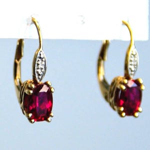 Puede incluir: Un par de pendientes dorados con cierre de palanca. Cada pendiente presenta una vibrante piedra preciosa roja oscura de corte ovalado. La parte superior de cada pendiente tiene un adorno en forma de hoja con pequeñas piedras transparentes.