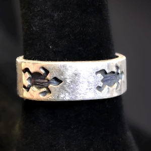 May include: A silver-toned ring with a textured finish. The ring features cut-out turtle designs around the band. The ring is displayed on a black surface.