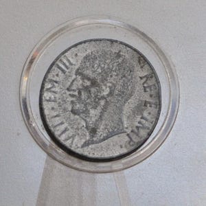 May include: A circular silver-toned coin encased in a clear rectangular plastic protective holder. The coin features a profile portrait of a person facing left, with the text "VITT EM III" on the left and "RE E IMP" on the right side of the face.