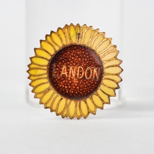 May include: A circular sunflower-shaped brooch featuring yellow enamel petals and a textured dark brown center. The name LANDON is embossed in capital letters across the center of the flower. The metal edges of the petals are outlined in gold.