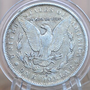 May include: A silver United States one dollar coin with an eagle design. The coin has the words "United States of America" and "In God We Trust" inscribed on it.