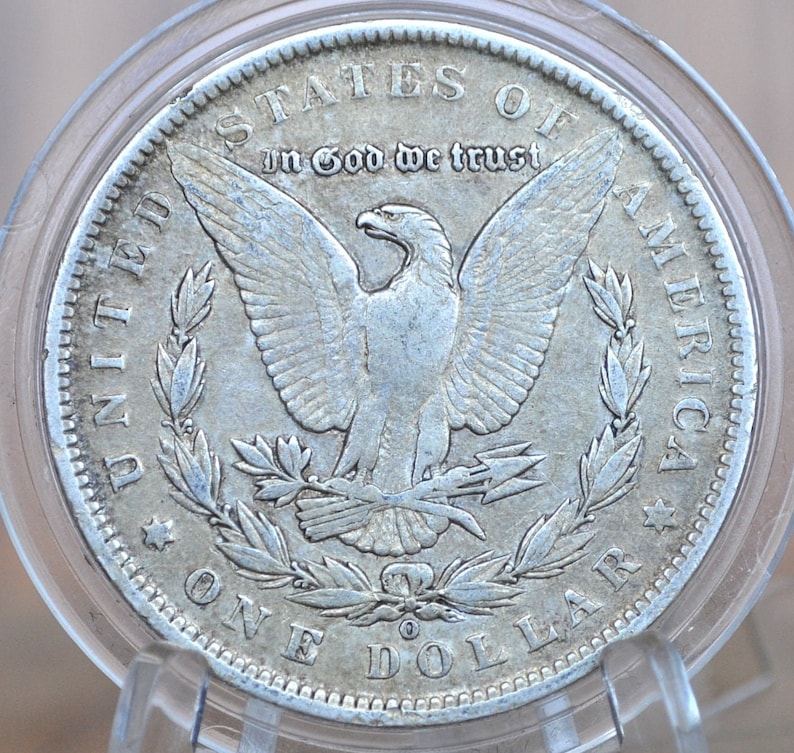 May include: A silver United States one dollar coin with an eagle design. The coin has the words "United States of America" and "In God We Trust" inscribed on it.