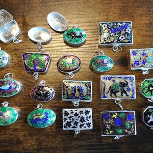 Puede incluir: Una colección de colgantes y pequeñas cajas plateadas ornamentadas con intrincados diseños de esmalte. Los colgantes presentan varios motivos de animales y flores en colores vibrantes como verde, azul y rojo. Las cajas tienen tapas con bisagras.