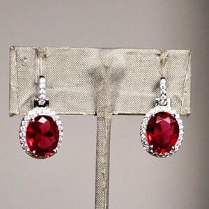 May include: A pair of elegant earrings featuring large, oval-cut, deep red gemstones. Each gemstone is surrounded by a halo of small, sparkling white stones, set in a silver-colored metal. The earrings are displayed on a neutral-toned background.