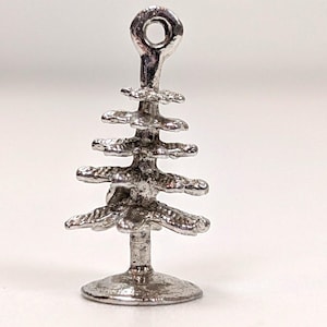 May include: A silver-colored Christmas tree charm with a loop at the top for hanging. The tree has multiple tiers of branches and a circular base. The charm is made of a shiny metal.