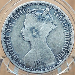May include: A silver coin with a portrait of Queen Victoria on the obverse. The coin has the inscription "Victoria D.G. Britt. Reg. F.D.  " around the edge.