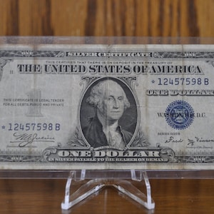 May include: A vintage one dollar silver certificate featuring a portrait of George Washington. The bill has a blue seal and the serial number 12457598 B. The text "THE UNITED STATES OF AMERICA" is printed in bold at the top of the bill.