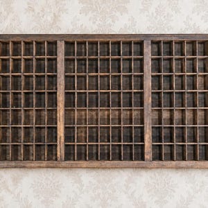 May include: A rectangular dark brown wooden printer's tray featuring a grid of numerous small, square compartments arranged in three distinct vertical sections. The wooden frame shows a weathered texture with visible grain and natural wood tones.