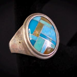 May include: Silver ring with an oval turquoise and brown mosaic inlay. The ring has a wide band and a polished finish. The mosaic design features geometric shapes in shades of blue, turquoise, and brown. A unique piece of jewelry.