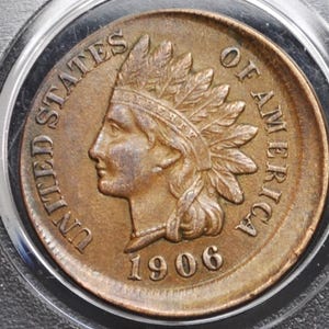 May include: A copper-colored 1906 Indian Head penny coin, encased in a protective holder. The coin features a profile of a Native American in a feathered headdress and the words "UNITED STATES OF AMERICA".