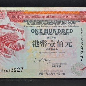 May include: A Hong Kong 100 dollar banknote with a lion's head on the left. The note has text in English and Chinese, with the number 100 in the top right. Serial number is EW633927.
