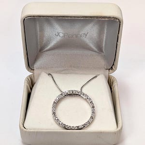 May include: A silver pendant necklace with a circle of small, clear stones, presented in an open, off-white jewellery box. The box has a satin lining and the brand name "JCPenney" is visible inside. The necklace is on a silver chain.
