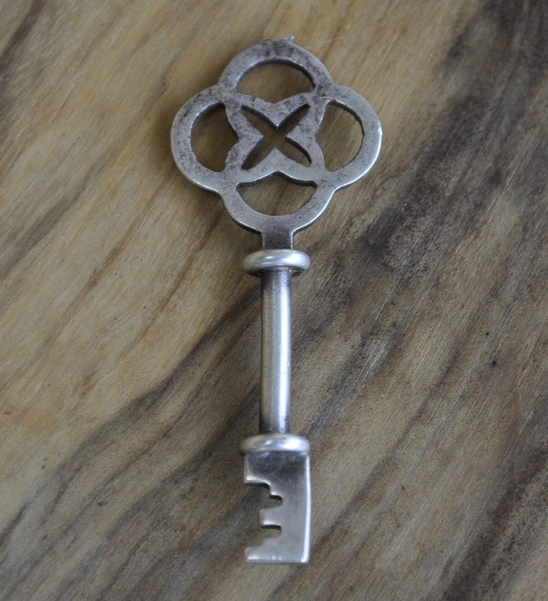May include: A silver key with a decorative, intricate, four-leaf clover design. The key has a long, straight shaft and a simple, rectangular blade.