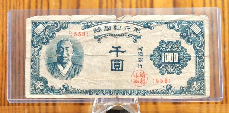 May include: A vintage Japanese 1000 Yen banknote with a blue and white design. The note features a portrait of a man in traditional clothing and the text "1000 Yen" in the centre. The note is in good condition and has a serial number of "558".