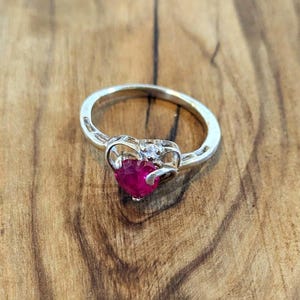May include: A silver ring with a heart-shaped, deep red gemstone as the centerpiece. The ring has a delicate design with a small, clear stone above the heart. The ring is set against a wooden background.
