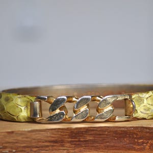 Könnte beinhalten: Ein gold-silbernes Armband mit Kettenglied-Design. Das Armband hat ein gelbes Schlangenmuster. Das Armband liegt auf einer Holzoberfläche.