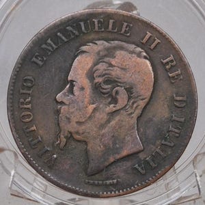 Puede incluir: Moneda de color cobre, dentro de un soporte de plástico transparente. La moneda muestra el perfil de un hombre con barba y la inscripción "VITTORIO EMANUELE II RE D'ITALIA" en el borde. Probablemente una moneda de colección antigua.