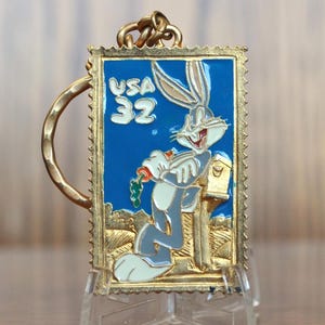 May include: Gold-colored metal keychain featuring Bugs Bunny holding a carrot and standing next to a mailbox. The keychain is shaped like a postage stamp with the text "USA 32" on it.