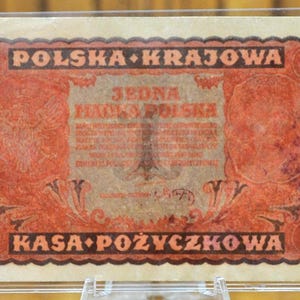 Puede incluir: Un billete de banco polaco vintage con un esquema de color rojo y blanco. El billete presenta un águila grande con una corona en la cabeza, y el texto "Polska Krajowa" y "Jedna Marka Polska" en letras negras. El billete también tiene el número "1" impreso en él.