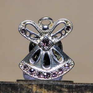 May include: Silver-toned angel pin with a halo and open-winged design. The angel's wings and dress are adorned with small, purple gemstones. The pin is set on a dark background.