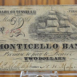Könnte beinhalten: Ein alter Zwei-Dollar-Banknote der Monticello Bank in Charlottesville, Virginia. Der Schein zeigt ein Porträt einer Frau, ein Schiff und den Text "Promise to pay to Bearer Two Dollars".