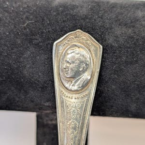 May include: A silver spoon with an engraved portrait of a man in an oval frame. The name "THOMAS MEIGHAN" is inscribed below the portrait. The spoon's handle features ornate floral designs.
