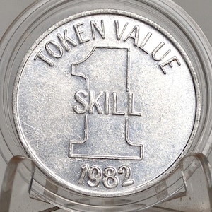 Könnte beinhalten: Ein silberner Token mit den Worten "TOKEN VALUE" oben. Die Zahl "1" befindet sich in der Mitte, darunter "SKILL" und unten "1982". Der Token ist in einem transparenten Kunststoffhalter eingeschlossen.