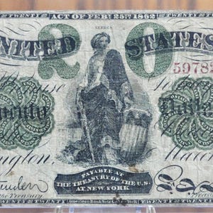 Puede incluir: Un billete de banco estadounidense de veinte dólares de 1862, con un retrato de una mujer con un bastón y un escudo, con el texto "TWENTY DOLLARS" en los lados izquierdo y derecho del billete, y "UNITED STATES" y "20" en la parte superior.